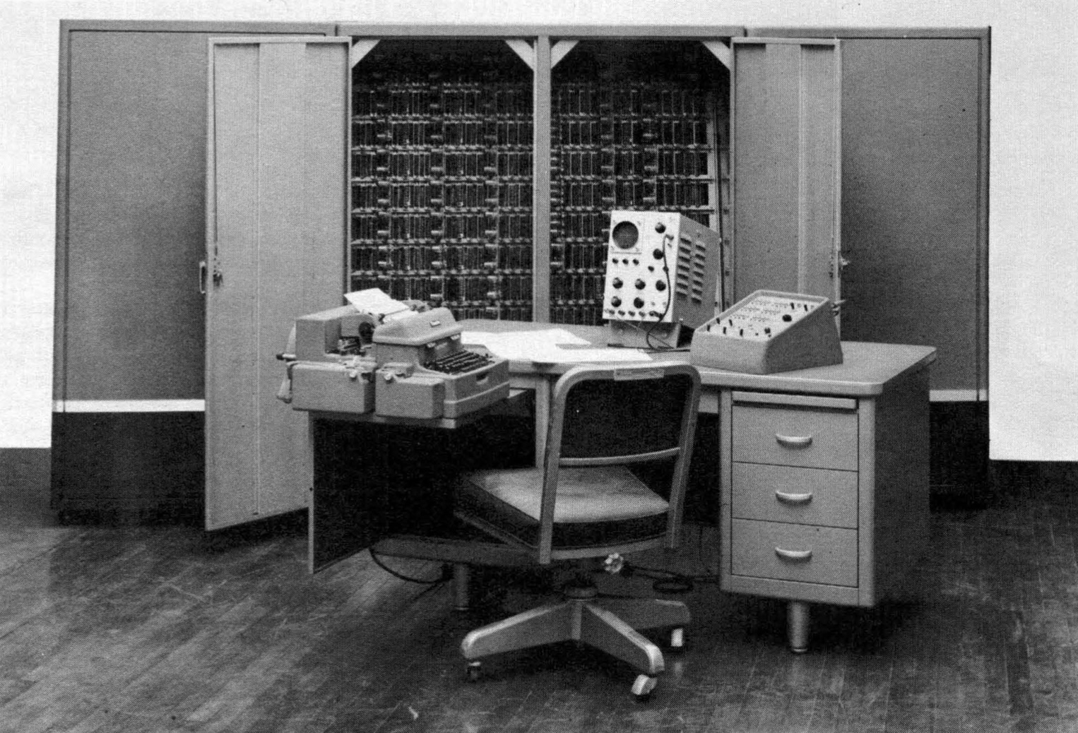 A photograph of a bare-bones ALWAC III-E. The operator desk in the foreground holds the
                         Flexowriter used for input and out, the control console, and an oscilloscope. In the background
                         are four cabinets housing storage and processor units.