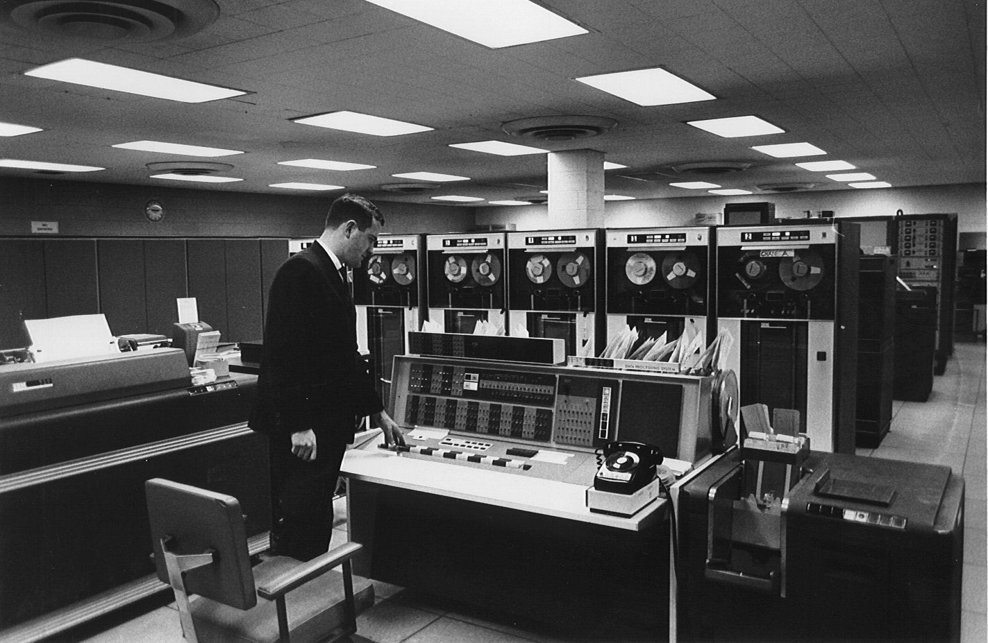 A man standing in front of the console of an IBM 7094 computer. There are six
                                 tape drives behind the console, a card punch/reader to his right, and a large
                                 line printer to his left.